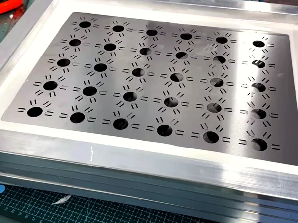 Framed SMT stencil with round laser-cut apertures resting on a green cutting mat